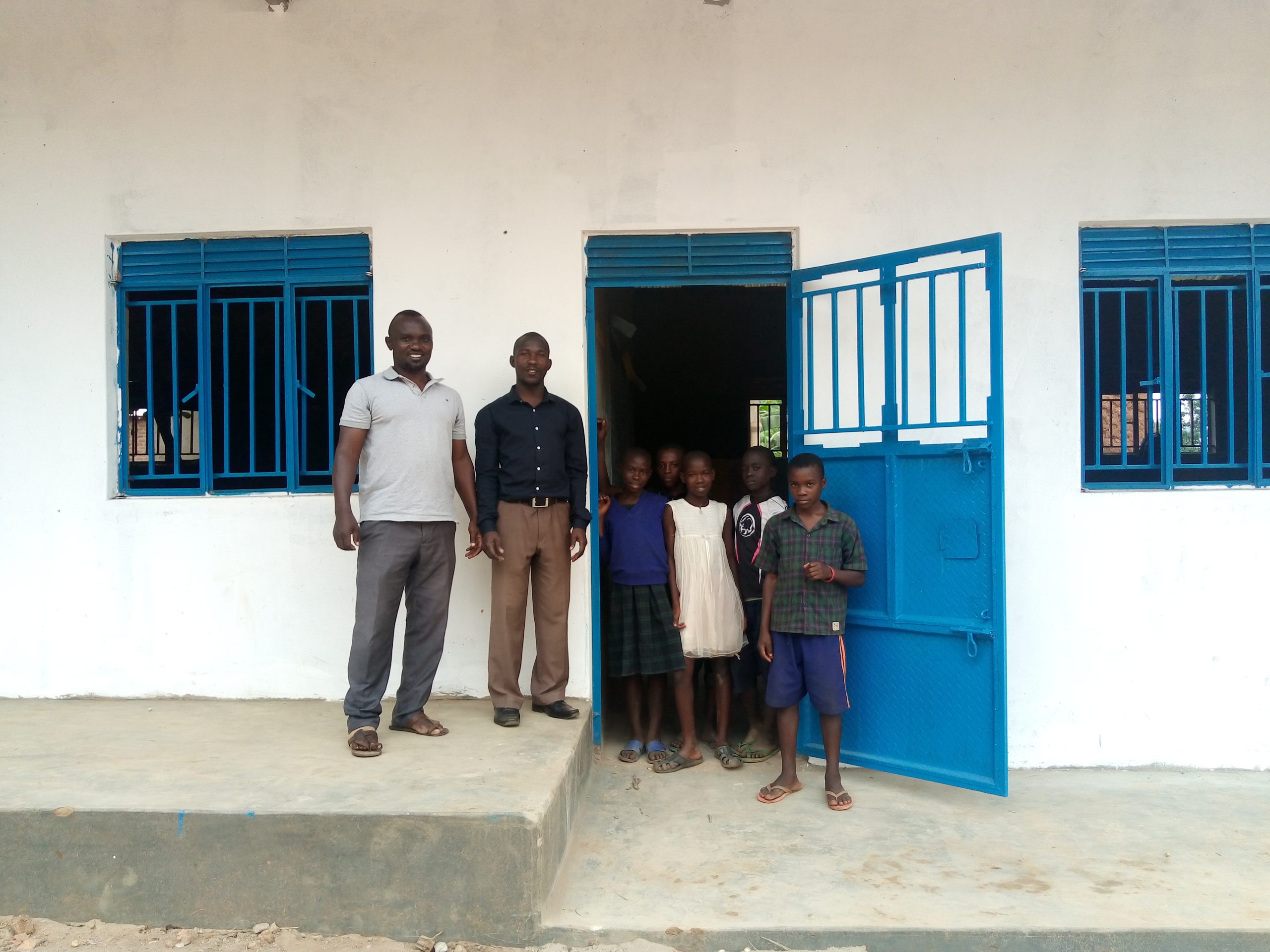 De directeur van een school in Oeganda staat voor een nieuw schoolgebouw met leerlingen. =