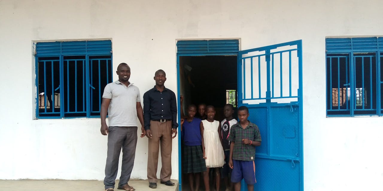 De directeur van een school in Oeganda staat voor een nieuw schoolgebouw met leerlingen. =