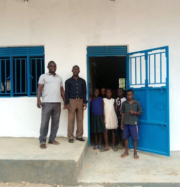 De directeur van een school in Oeganda staat voor een nieuw schoolgebouw met leerlingen. =