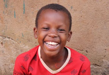 Een jongetje met een rood shirt lacht in de camera in Oeganda.