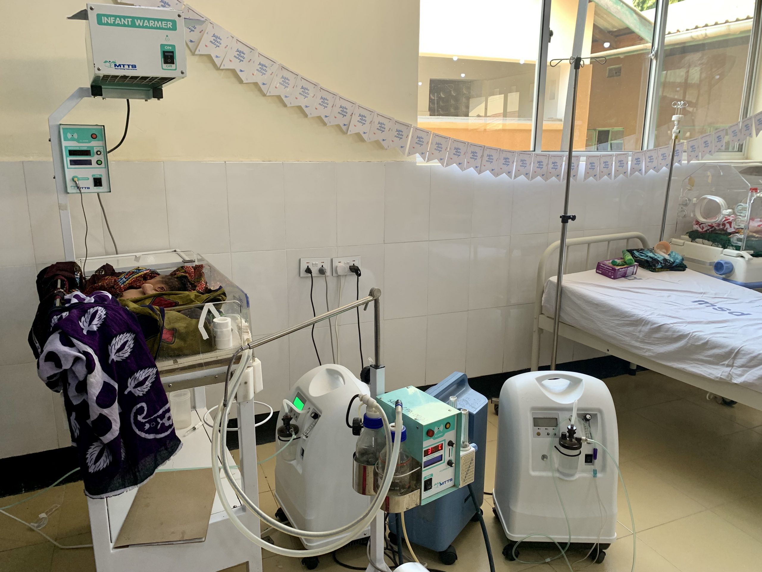 Een baby in een 'infant warmer'. Op de achtergrond een slinger met Wilde Ganzen kaartjes.