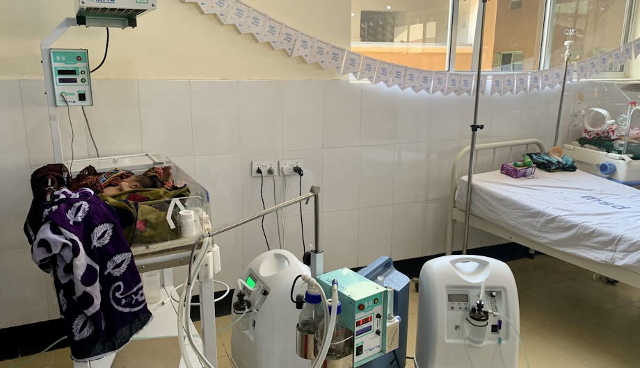 Een baby in een 'infant warmer'. Op de achtergrond een slinger met Wilde Ganzen kaartjes.