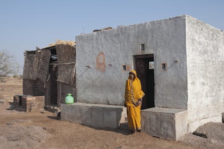 Een vrouw in een geel gewaad staat voor een betonnen huis in India.